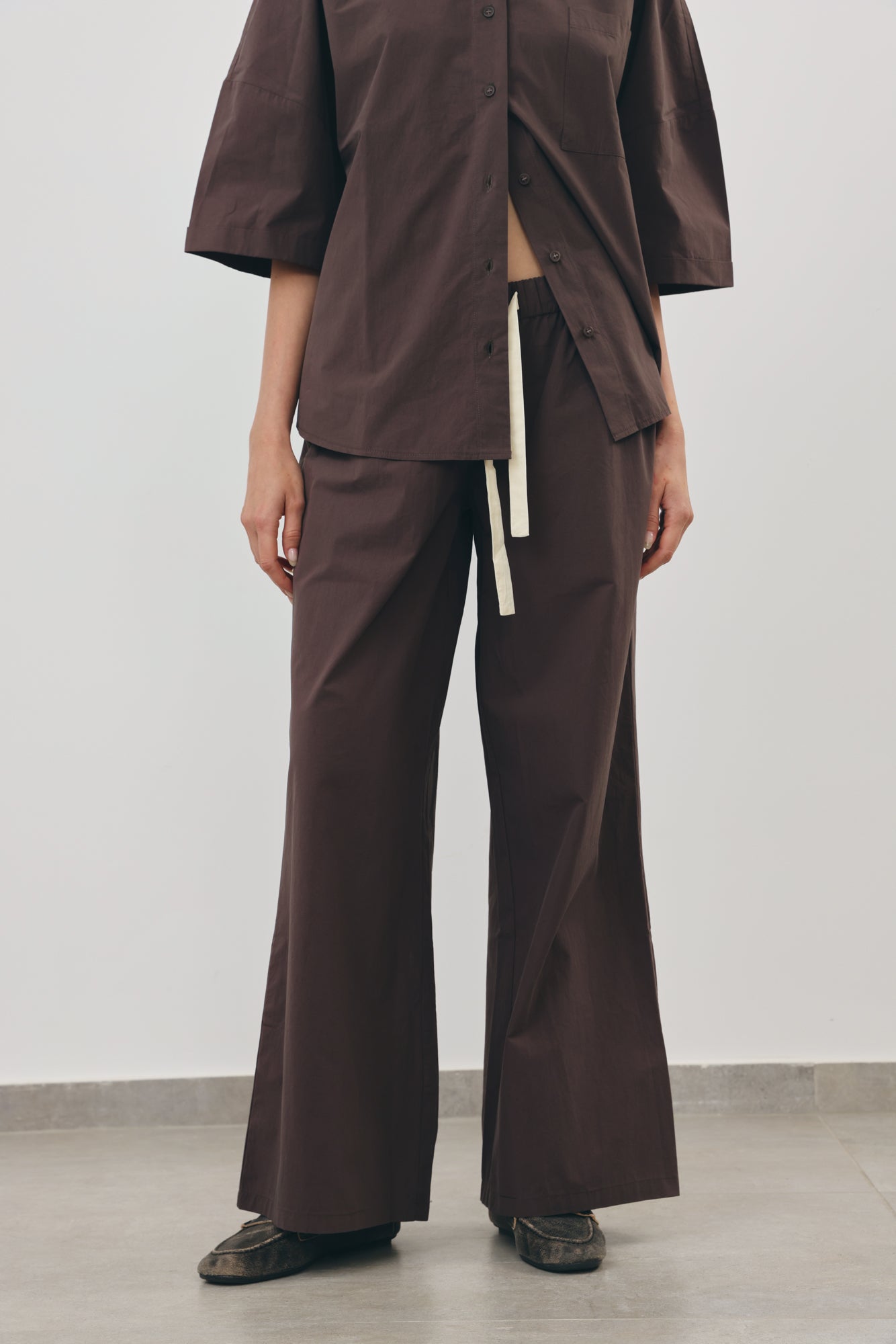 A model wearing a matching dark brown two-piece outfit consisting of a short-sleeve button-front top and wide-leg trousers with a cream drawstring, paired with dark loafers against a minimalist studio background.