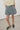 A person wearing high-waisted, wide-leg denim shorts in a faded light blue wash, paired with a cream shirt and dark loafers, standing against a plain light gray wall and floor.