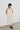 A woman with short black hair stands against a white wall wearing a cream-colored linen slip dress with lace trim on the straps and square neckline, paired with dark loafers.
