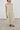 A woman modeling a saga midi dress with a smocked bodice, thin shoulder straps, and a gathered skirt, paired with brown suede flats against a neutral studio background.
