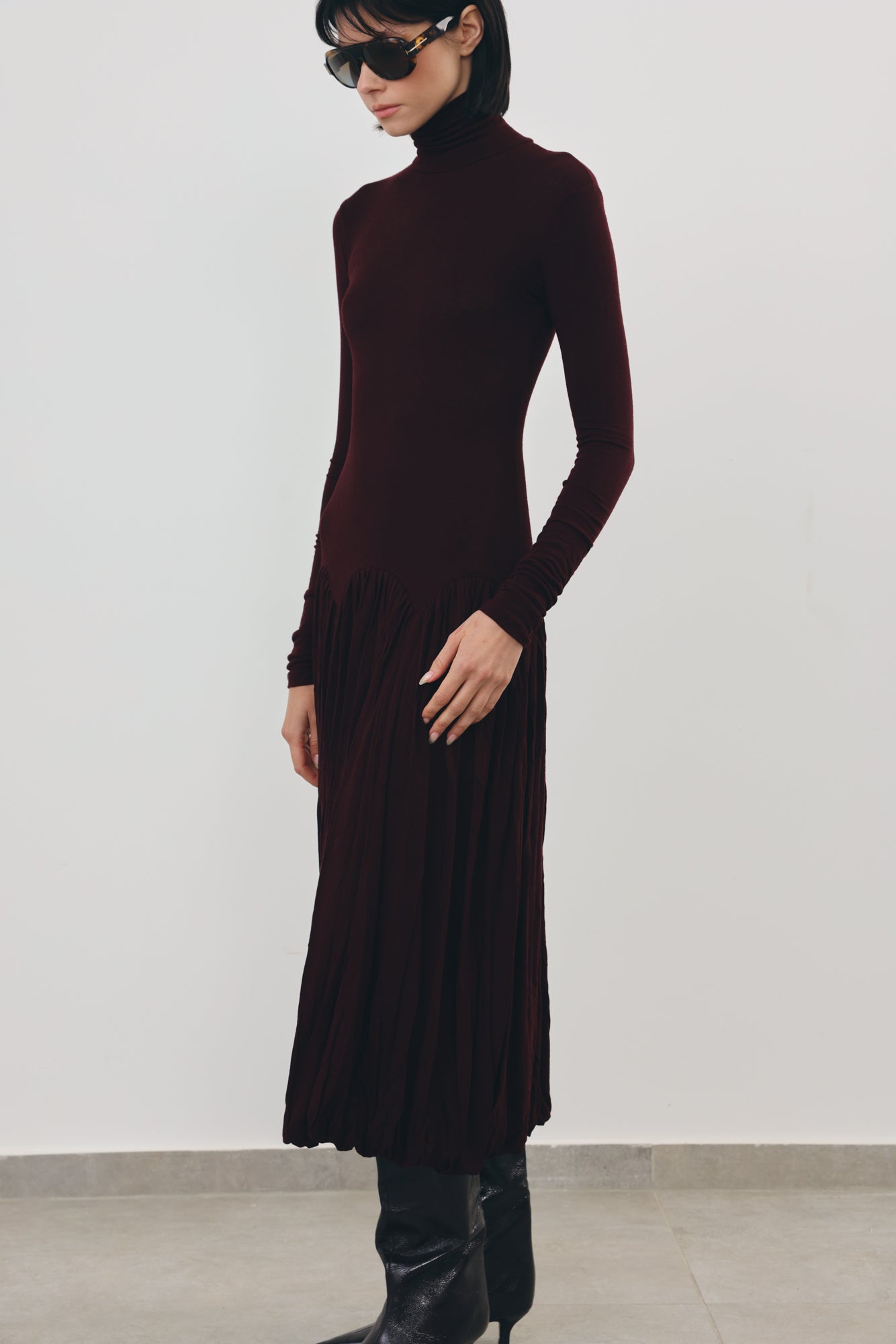 A model wearing a dark burgundy long-sleeve turtleneck dress with a pleated skirt, styled with black boots and sunglasses against a plain light gray wall.